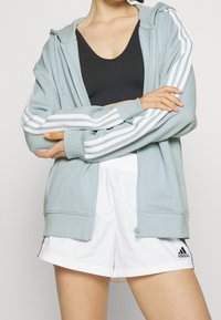 Světlé modrá mikina na zip s bílými pruhovanými akcenty na rukávech, doplněná bíl ými šortkami. Hladká látka a volný střih.