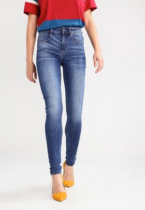 Person iført falmede blå skinny jeans, rød og blå t-shirt og orange spidse hæle, stående mod en neutral baggrund.