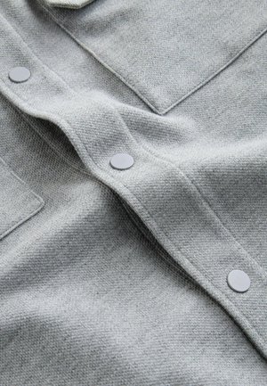 Gråt vævet stof med en tekstureret overflade, der har flade grå knapper og en lommedetalje med klap. Materialet er blødt og let elastisk.