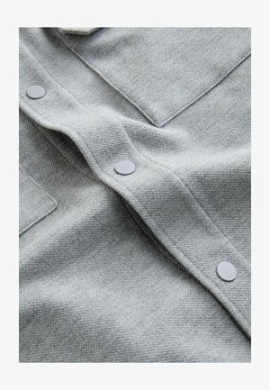 Grauer gewebter Stoff mit strukturierter Oberfläche, versehen mit flachen grauen Knöpfen und einer Klappentaschen-Details. Das Material ist weich und leicht dehnbar.