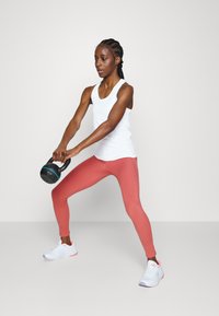 Balta sporta bez piedurknēm t-krekls, rozā legingi un baltas sneakers. Individuāls tur melnu kettlebellu, nostājies dinamiski vingrošanas pozā.