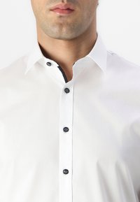 Hvit skjorte med klassisk krage, som har svarte knapper og kontrasterende kant langs plaggstykket. Glatt, ensfarget stoffstruktur.