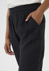 I pantaloni neri in lino presentano una vita ampia con pieghe e tasche laterali. Il tessuto ha una finitura morbida e testurizzata.