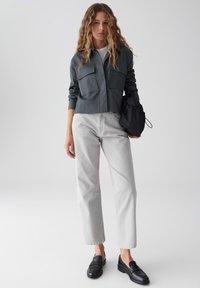 Camicia grigia scura con bottoni e due tasche sul petto, abbinata a pantaloni grigi chiari dritti. Mocassini neri e una borsa nera completano il look.