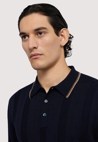 Námořnické žebrované polo tričko s kontrastním béžovým límcem a třemi modrými knoflíky. Navrženo s vertikálními pruhy a jemnou texturou.