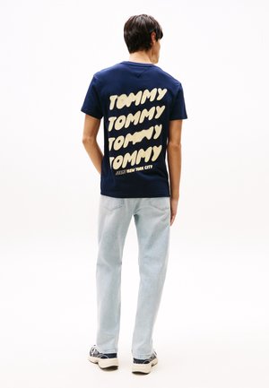 Homme portant un t-shirt bleu marine avec un grand texte "TOMMY" répété et un jean bleu clair, debout de dos sur fond blanc.