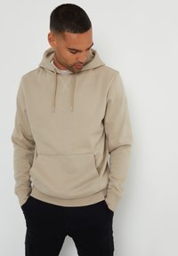 Sudadera con capucha beige de tejido suave, con un bolsillo frontal, cordón ajustable y puños acanalados. Combinada con pantalones negros.