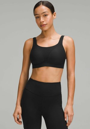 Sujeción deportiva negra con tirantes anchos, diseño sin costuras, copas acolchadas y detalles perforados en la parte delantera. Combinada con pantalones leggings negros de cintura alta.