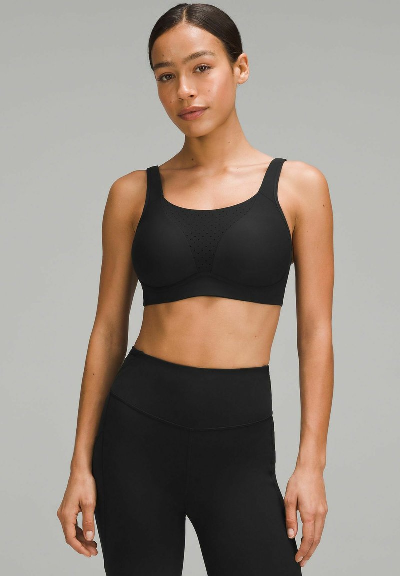 Sujeción deportiva negra con tirantes anchos, diseño sin costuras, copas acolchadas y detalles perforados en la parte delantera. Combinada con pantalones leggings negros de cintura alta.
