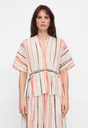 Kobieta w kremowej bluzce z krótkim rękawem i spódnicy z pionowymi paskami w odcieniach pomarańczowym i brązowym na jednolitym białym tle.