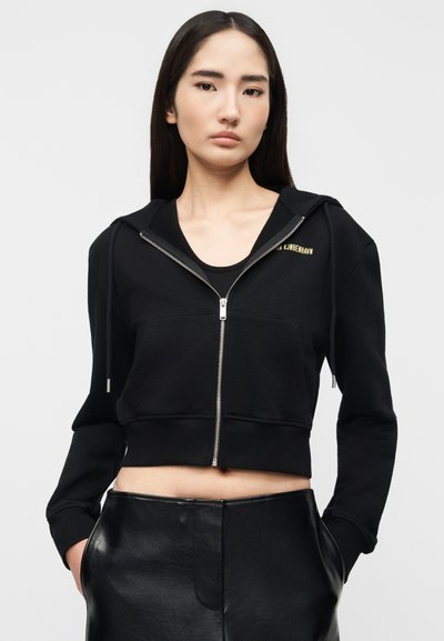 Νεαρή γυναίκα που φοράει μαύρο κοντό φούτερ με φερμουάρ και χρυσά γράμματα κοντά στον γιακά, καθώς και μαύρο δερμάτινο παντελόνι, με τα χέρια στις τσέπες.