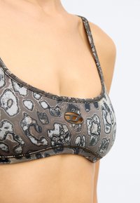 Στηθόδεσμος με λεοπάρ σχέδιο σε αποχρώσεις καφέ και γκρι, διαθέτοντας στρογγυλεμένο σχήμα, ρυθμιζόμενα λουράκια και μια μικρή λογότυπο λεπτομέρεια στο μπροστινό μέρος.