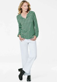 Grüne gemusterte Bluse mit V-Ausschnitt und langen Ärmeln, kombiniert mit weißen Hosen und schwarzen Stiefeletten mit Absatz. Glattes Material, moderner Schnitt.