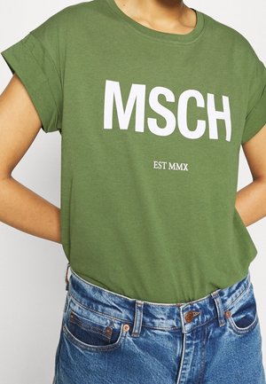 Personne portant un t-shirt vert avec le texte blanc « MSCH » et « EST MMX » associé à un jean en denim bleu.