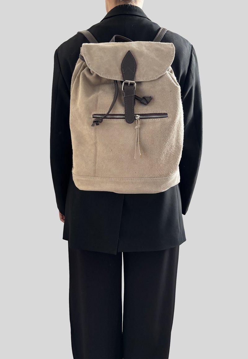 Béžový látkový batoh se stahovací šňůrkou, koženými detaily, zipovou přední kapsou a klopou nahoře. Obsahuje nastavitelné popruhy.