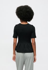 Kobieta z naturalnie kręconymi włosami, ubrana w dopasowaną, czarną bluzkę z krótkim rękawem oraz jasnoszare spodnie w prążek, stojąca na jednolitym tle.