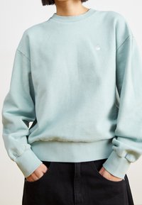 Persona vistiendo un suéter azul claro de cuello redondo con mangas largas y pantalones negros, con las manos en los bolsillos, frente a un fondo liso.