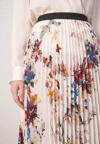 Jupe blanche plissée avec imprimé floral multicolore, ceinture noire avec texte embossé, associée à un chemisier crème à manches longues.