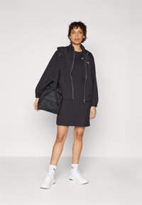 Veste bomber noire zippée avec des poignets côtelés, associée à une robe noire. Des chaussures de sport blanches et un sac à épaule noir complètent le look.