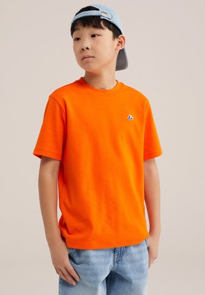 Niño con una camiseta naranja brillante de manga corta con un pequeño logo y una gorra azul claro hacia atrás, de pie frente a un fondo liso.