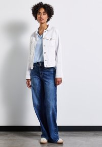 Biała dżinsowa kurtka z dwiema kieszeniami na piersi, jasnoniebieska koszulka z dekoltem w serek, ciemnoniebieskie szerokie dżinsy i beżowe trampki na jednolitym tle.