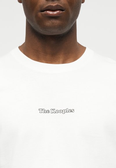 Κοντινό πλάνο ενός ατόμου που φοράει λευκό πουκάμισο με στρογγυλή λαιμόκοψη και την επιγραφή «The Kooples» σε μαύρο στο στήθος.