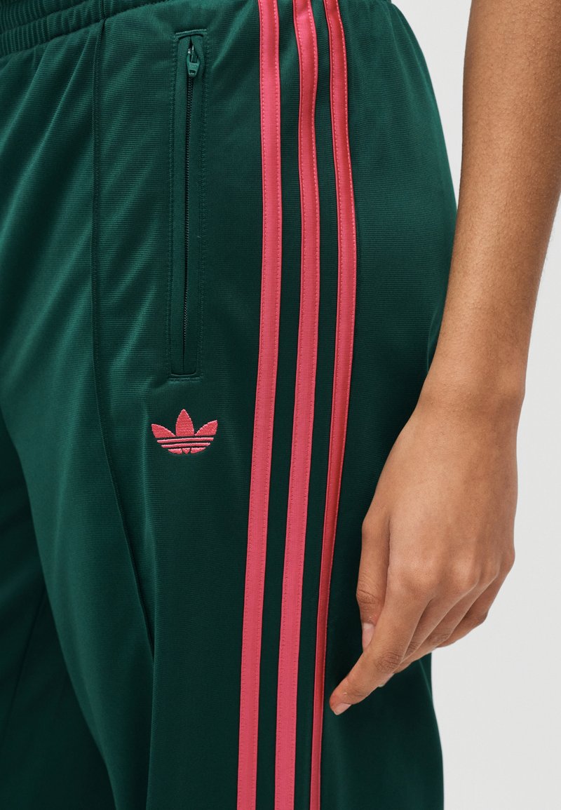 Pantalon de sport vert avec des bandes latérales roses et un logo Adidas rose, doté d'une poche zippée près de la taille et d'un bras visible à côté.