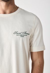 Cream-colored short-sleeve t-shirt, made of cotton, featuring a green embroidered logo on the left chest area with detailed text.
