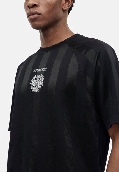 Νέος άντρας που φοράει μαύρο ριγέ T-shirt με λευκό έμβλημα και το κείμενο «HAN KJØBENHAVN» στο στήθος, στέκεται μπροστά σε απλό φόντο.