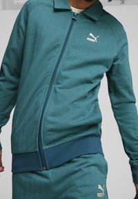 Teal zip-up jakke med herringbone tekstur, to lommer og ribbet manchetter og kant; parret med matchende bukser. Puma-logoen vises.