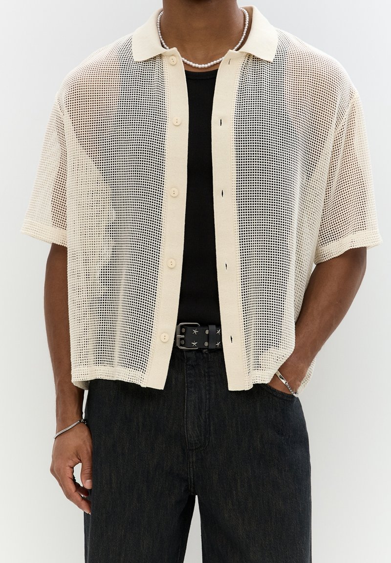 Mies, joka yllään on vaalean beige lyhythihainen verkko-tyylinen nappipaita mustan toppatakin päällä, mustat housut, joissa on tähtiä oleva vyö, ja hopeiset korut.