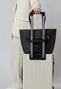 Schwarze Leder-Tragetasche mit einer Reißverschlusstasche, die auf einem cremefarbenen Hartschalenkoffer mit vertikalen Rillen und einem ausziehbaren Griff liegt.