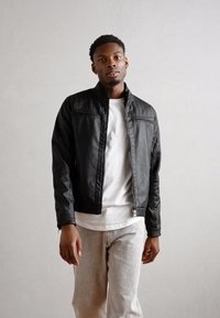 Jachetă din piele neagră cu guler înalt, detalii de cusături diagonale, închidere cu fermoar în față și o textură netedă. Pur tată peste o cămașă albă și pantaloni deschiși la culoare.