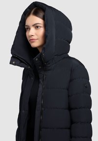 Tamno plava puffer jakna s velikom kapuljačom, koja ima patentni zatvarač i kopče na gumbi, teksturirani kviltani dizajn i logo akcent na rukavu.