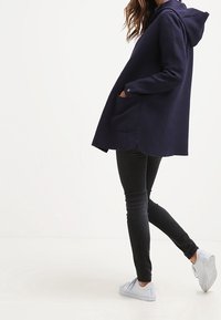 Marineblå hættejakke med frontlommer, lange ærmer og afrundet kant. Båret over sorte skinny jeans og hvide sneakers. Glat tekstur.