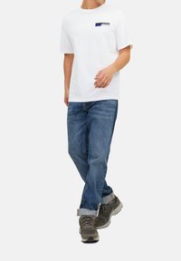 Hvid t-shirt med korte ærmer og blå og sort grafiktryk på venstre bryst, parret med blå denim jeans og grå sneakers.