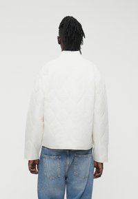 Balta dygsniuota striukė su aukštu apykakliu ir ilgomis rankovėmis, turinti tekstūruotą raštą. Derinama su mėlynais džinsais.