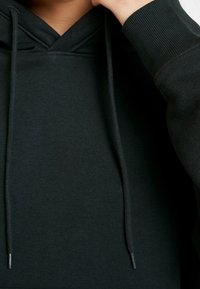 Černá mikina s designem ve tvaru V, vyrobená z měkké tkaniny. Má šňůrky na stažení a žebrované manžety pro těsné přilnutí.