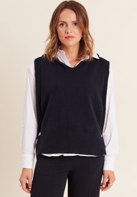Pull sans manches en tricot bleu marine avec ourlet côtelé, présentant un col en V et des détails de boutons sur les côtés, superposé sur une chemise blanche.