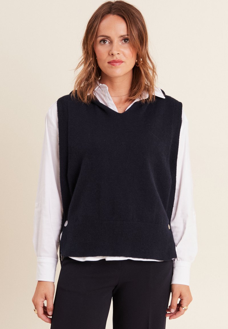 Pull sans manches en tricot bleu marine avec ourlet côtelé, présentant un col en V et des détails de boutons sur les côtés, superposé sur une chemise blanche.