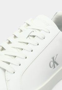 Λευκό δερμάτινο sneaker με ίσιες κορδόνες, υφή λεπτομέρεια και γκρι λογότυπο "cK" στο πλάι. Μινιμαλιστικός σχεδιασμός, λεία επιφάνεια και στρογγυλή μύτη.