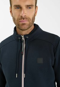 Giacca a zip navy in morbido tessuto con colletto alto. Presenta una zip a contrasto e una patch con logo sul petto. Design pulito e moderno.