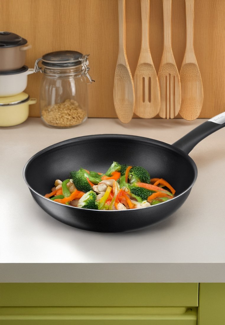 Zwarte anti-aanbak koekenpan met roergebakken groenten, waaronder broccoli, paprika en gesneden kip, op een lichte aanrecht.