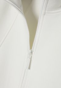 Giacca bianca con zip, colletto a coste, tessuto liscio e cerniera in plastica visibile. Primo piano mostra il dettagli della cerniera e delle cuciture.