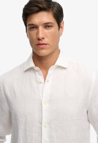 Hombre con cabello corto y oscuro que lleva una camisa de lino blanca sin abotonar sobre un fondo claro y liso.