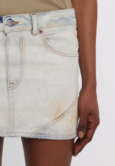 Fustă mini din denim deschis, cu un aspect decolorat, detalii cu cusături curbe și accente distorsionate. Include un nasture metalic și buzunare frontale.