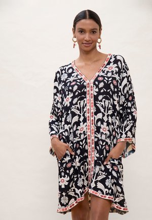 Mujer con vestido negro de flores con ribete coral, de pie con las manos en los bolsillos frente a un fondo neutro claro.