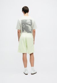 Tricou gri deschis cu un imprimeu grafic mare pe spate, asortat cu pantaloni scurți verzi deschis. Material neted, croială lejeră, adidași albi.
