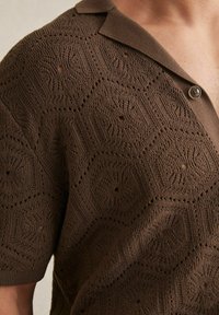 Polo din tricot de culoare maronie, cu un model în fagure, mâneci scurte și un guler moale. Are găuri decorative și un singur nasture.