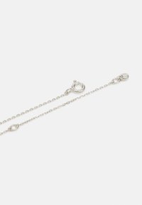 Sølv kædedesign med en sammenkoblet struktur, rund lås og et lille logo-tag i den ene ende. Glat tekstur og poleret finish.